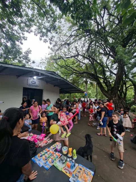 Familias y voluntarios reunidos alrededor de una mesa con juguetes y refrigerio al aire libre.