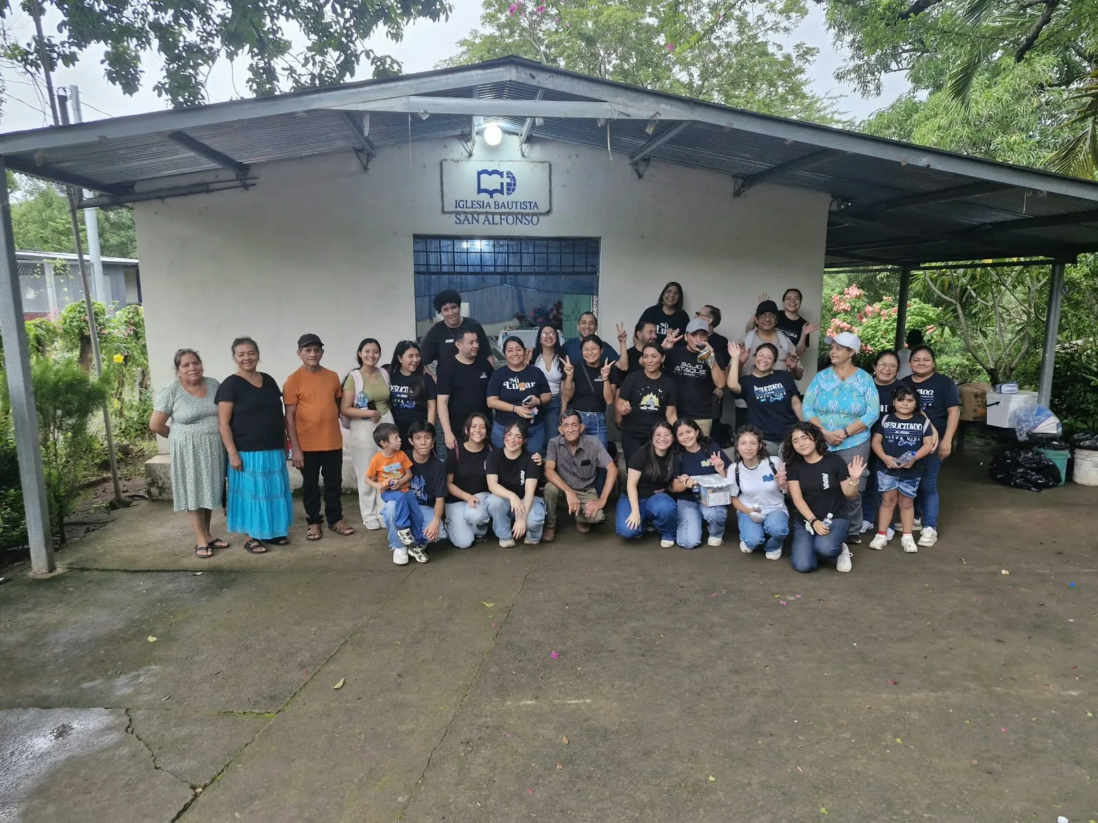 Participantes del viaje misionero reunidos frente a la fachada de la Iglesia Bautista San Alfonso, rodeados de árboles.